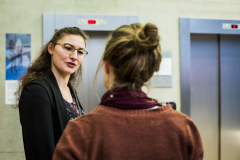 Zwei Frauen stehen vor zwei Aufzügen, eine im Vordergrund mit Dutt und Schal, eine links im Profil.