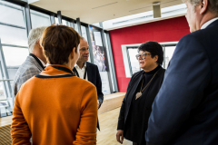 Fünf Personen stehen in einem hellen Raum mit großen Fenstern; eine trägt einen orangefarbenen Pullover.