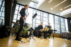 Musikgruppe mit Schlagzeug, E-Bass, Saxophon und Keyboard in einem hellen Raum mit großen Fenstern.
