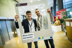 Zwei Männer halten einen großen Rahmen mit der Aufschrift „#BEACTIVE“ und „Move more, feel better.“