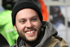 Person mit schwarzer Mütze und grauem Kapuzenpullover im Vordergrund; im Hintergrund zwei weitere Personen.