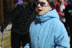 Person im hellblauen Mantel und mit violetten Handschuhen hält blaue und violette Bänder im Schnee.