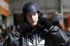 Person mit Helm, schwarzer Brille, Handschuhen und grauer Jacke im Vordergrund; weitere Personen im Hintergrund.