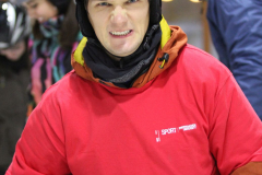 Drei Personen mit Helmen, im Vordergrund eine Person in rotem Shirt und brauner Jacke, Hände vor dem Bauch.