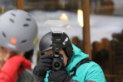 Person mit Helm und Kamera fotografiert in einer Halle; weitere Personen im Vordergrund unscharf.