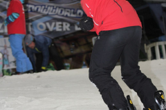 Snowboarder in roter Jacke und schwarzer Hose auf der Piste, im Hintergrund weitere Personen und ein Banner.
