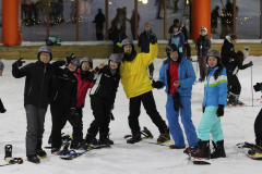 Mehrere Personen mit Snowboards und Helmen stehen auf einer Indoor-Skipiste.