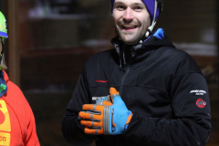Person mit schwarzem Helm, schwarzer Jacke und blauen-orangefarbenen Handschuhen; links teilweise Person in roter Jacke.
