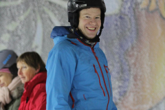 Person in blauer Skijacke, schwarzem Helm und Handschuhen vor gemalter Wand; zwei Personen links im Hintergrund.
