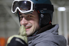 Nahaufnahme einer Person mit Skihelm, Skibrille und grauer Jacke; Gesichtszentrum verpixelt.
