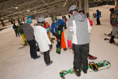Mehrere Personen mit Helmen stehen in einer Skihalle auf Schnee, einige halten Snowboards.