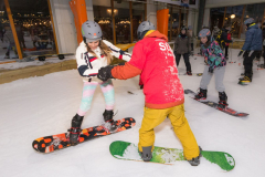 Mehrere Personen mit Helmen stehen auf Snowboards in einer Halle auf Schnee.