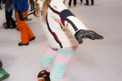 Person im Schneeanzug und Helm steht auf einem Snowboard in einer Halle, andere Personen im Hintergrund.