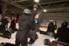 Snowboardkurs in einer Halle: mehrere Personen mit Helmen und Snowboards im Schnee, eine Person hilft einer anderen beim Aufstehen.