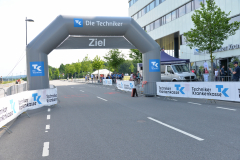 Zielbogen an einer gesperrten Straße mit Techniker-Krankenkasse-Bannern, Zuschauer und Gebäude rechts.