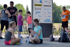 Menschen sitzen und stehen draußen neben einem großen Sponsoren-Schild; Fahrräder im Hintergrund.