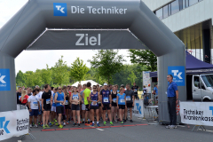 Läufergruppe vor einer grauen Ziel-Start-Arch mit „Die Techniker“ und „Ziel“ sowie Absperrungen und einem Transporter.