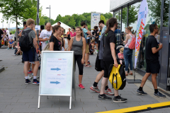 Menschen stehen vor einem Gebäude, ein Schild mit Pfeilen steht im Vordergrund, rechts ein buntes Banner.