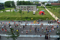 Viele Läuferinnen und Läufer auf einer abgesperrten Straße, daneben Zuschauer, Zelte und Gebäude.