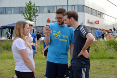 Drei Personen stehen auf einer Wiese; die mittlere hält ein Smartphone. Im Hintergrund ein Gebäude und weitere Menschen.