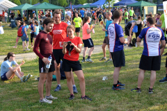 Menschen auf einer Wiese bei einer Sportveranstaltung mit Zelten im Hintergrund, einige halten Getränke und Snacks.