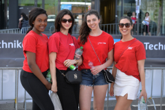 Vier Frauen in roten T-Shirts stehen vor einer Absperrung; zwei halten Flaschen, eine Tasche, eine Plastiktüte.