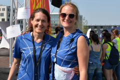 Zwei Personen in blauen Sportshirts stehen vor einer Bühne mit rotem Banner und weiterer Menschenmenge.