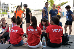 Mehrere Personen in Sportkleidung und roten STAFF-T-Shirts sitzen und stehen draußen auf einer breiten Promenade.