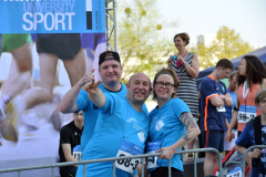 Drei Personen in blauen T-Shirts posieren vor einer Absperrung bei einer Sportveranstaltung.