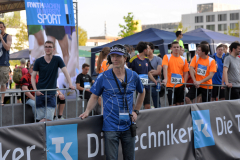 Person in blauem Shirt mit Headset vor Absperrung; dahinter mehrere Personen und Zelte.