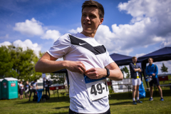 Mann im weißen Sportshirt mit Startnummer 491 steht im Freien, im Hintergrund Zelte, Bäume und andere Personen.