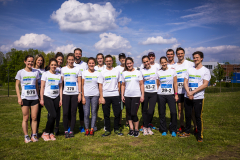 Gruppe von 14 Personen in weißen T-Shirts und Laufnummern steht auf einer Wiese unter blauem Himmel.