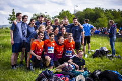 Gruppe junger Menschen im Gras; zwei tragen orange Trikots mit Startnummern, Taschen liegen vorne.