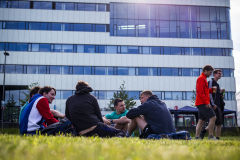 Mehrere Personen sitzen und stehen auf einer Wiese vor einem modernen Gebäudekomplex.