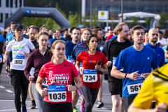 Läufergruppe mit Startnummern auf einer Straße vor Gebäuden und einem Startbogen.