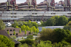 Großes Gebäude mit Rohren im Hintergrund, davor Bäume, Häuser und unten drei laufende Personen.