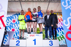Siegerpodest mit fünf Personen, drei auf dem Podest, zwei daneben, vor bunten Sport-Bannern.