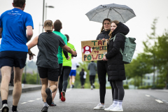 Menschen laufen auf einer Straße; zwei Personen stehen mit Regenschirm und halten ein Schild mit Text.