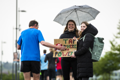 Drei Personen auf einer Straße; zwei halten einen Regenschirm und ein Schild mit der Aufschrift „TAP HERE TO POWER“.