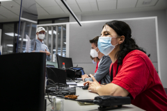 Drei Personen mit Masken sitzen an Laptops in einem Besprechungsraum; eine Person steht im Hintergrund.