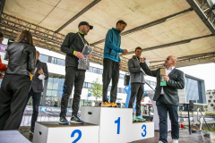 Männer auf einem Siegerpodest mit den Zahlen 1, 2 und 3, teils mit Pokalen und Medaillen unter einem Zelt.