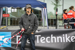 Mann mit Kopfhörern steht vor einem Banner, hält rot-weißes Absperrband; im Hintergrund zwei Personen in Warnkleidung.