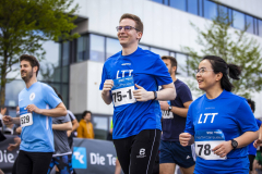 Mehrere Läufer in blauen T-Shirts mit Startnummern vor einem modernen Gebäude.