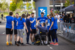 Gruppe von Läufern in blauen Shirts mit einer Person im Rollstuhl auf einer abgesperrten Straße.