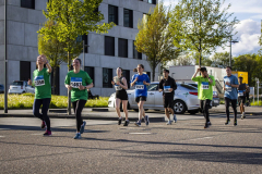 Läufergruppe mit Startnummern läuft auf einer Straße vor einem Gebäude und geparkten Autos.