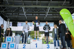 Siegerehrung auf einem Podest: drei Personen mit Medaillen und Urkunden, daneben weitere Personen und ein grünes Banner.