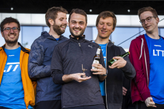 Fünf Männer stehen nebeneinander, drei halten Flaschen und einen kleinen Pokal.