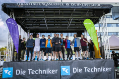 Gruppe von Männern auf einer Bühne, flankiert von zwei großen Bannern, vor Gebäuden und Himmel.