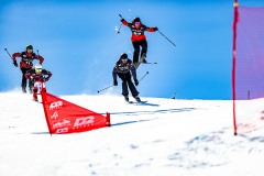 Vier Skifahrer springen über eine verschneite Rennstrecke mit roten Toren.