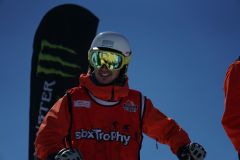 Snowboarder in roter Ausrüstung vor blauem Himmel mit großem Snowboard im Hintergrund.